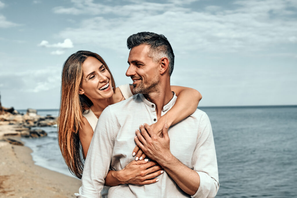 Woman holding her husband from behind standing at a beach on a sunny day. Romantic couple on standing near the sea together.